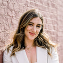 Woman with long hair and a white blazer standing against a pink brick wall.